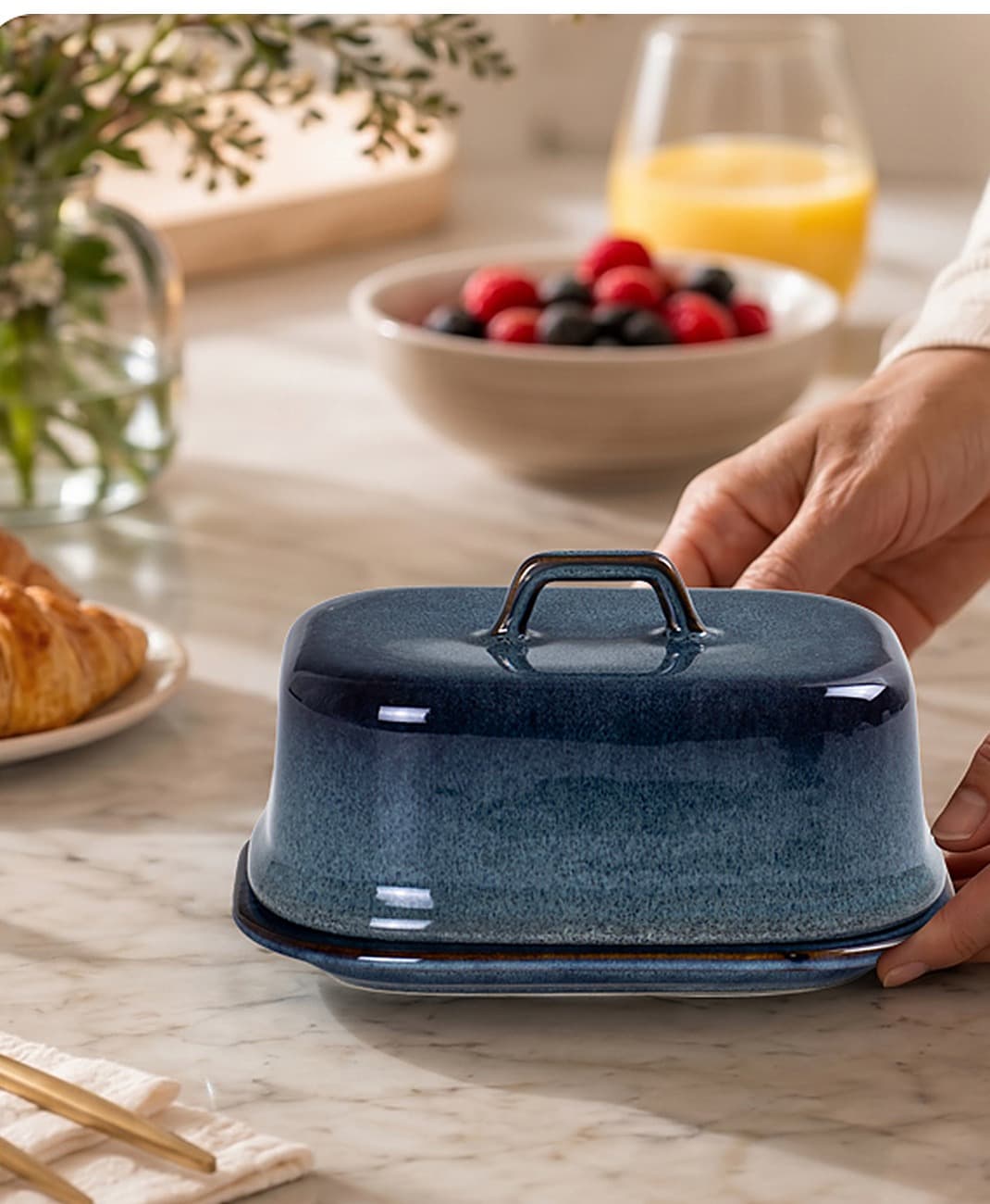 Unas manos colocan la mantequillera azul en una mesa de desayuno con mármol